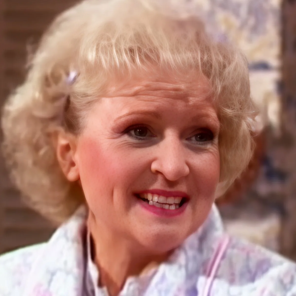 A smiling woman with blonde, curly hair wearing a light-colored, patterned top.