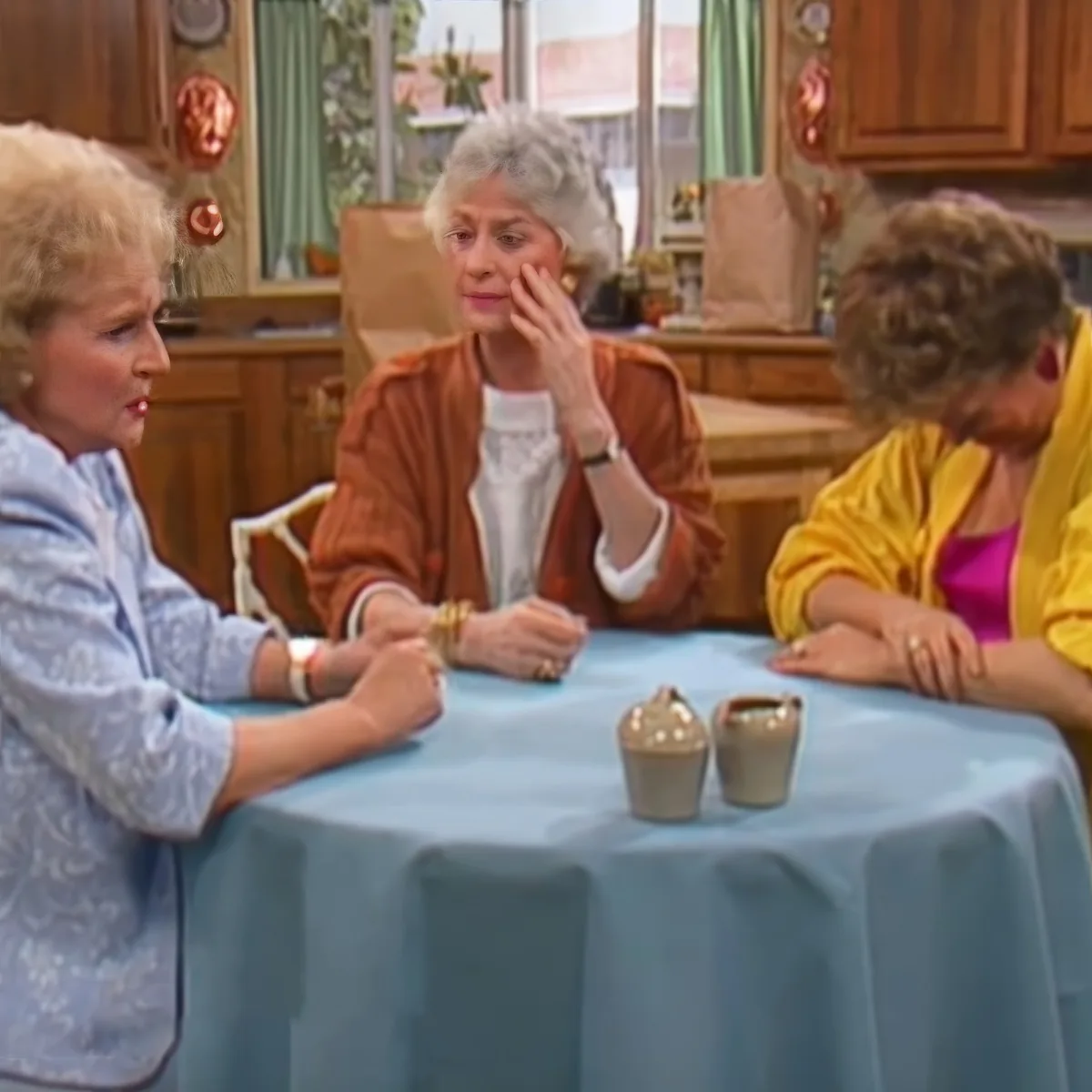 Three women sit at a table with a blue tablecloth, engaged in conversation in a kitchen setting.