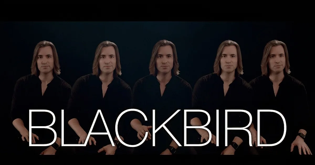A person with long hair and a black shirt gestures with their right hand against a dark background.