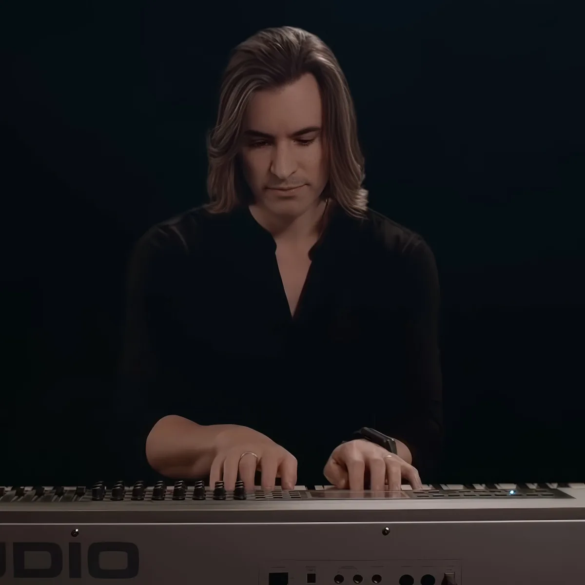 A person with long hair plays a keyboard in a dimly lit setting.