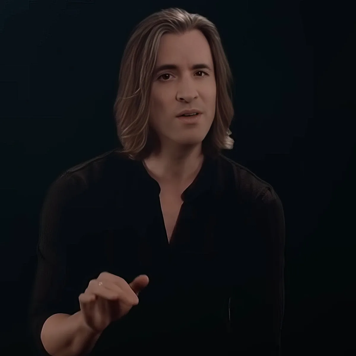 A person with long hair, wearing a black shirt, gestures with their hands while speaking against a dark background.