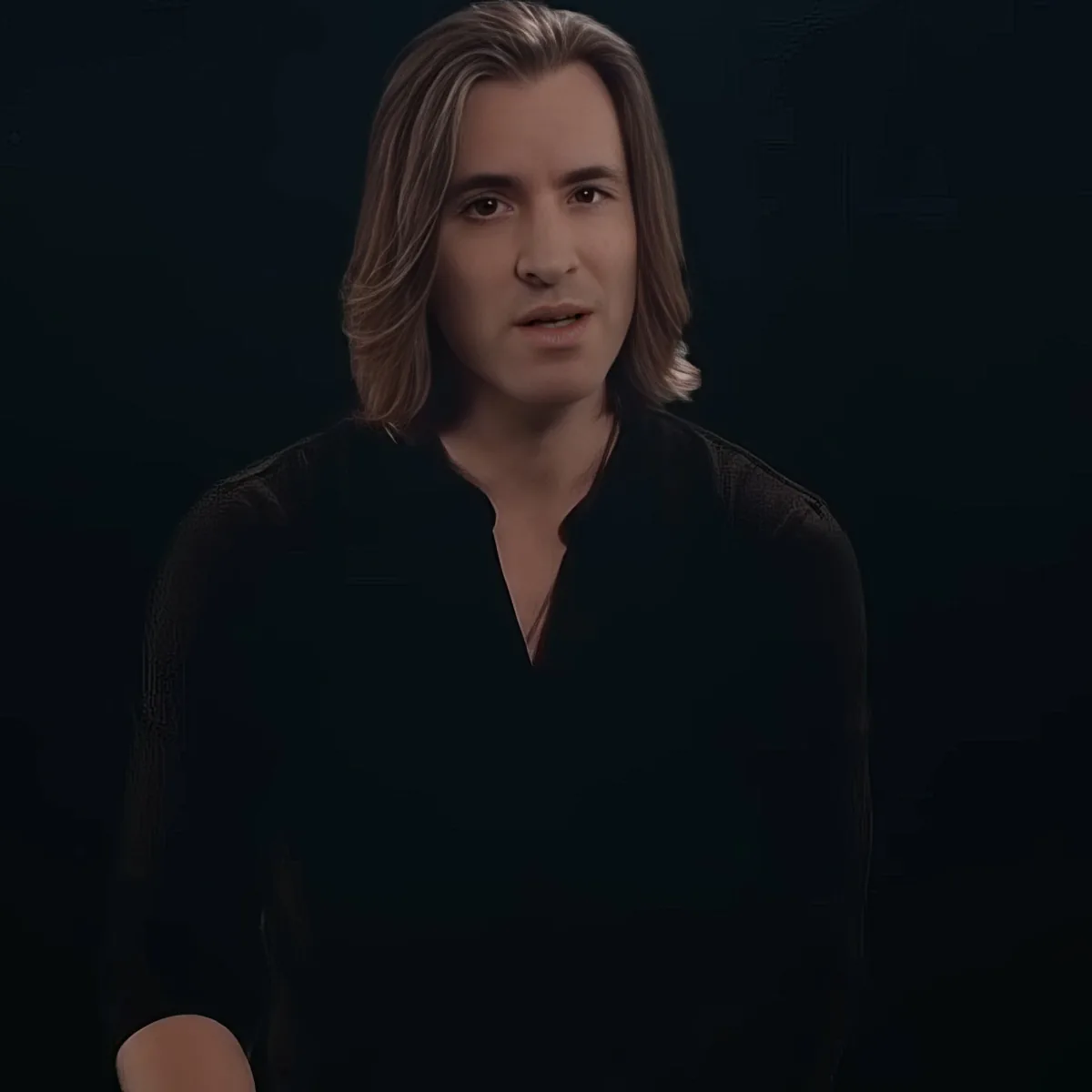 A person with long hair, wearing a black shirt, stands against a dark background.