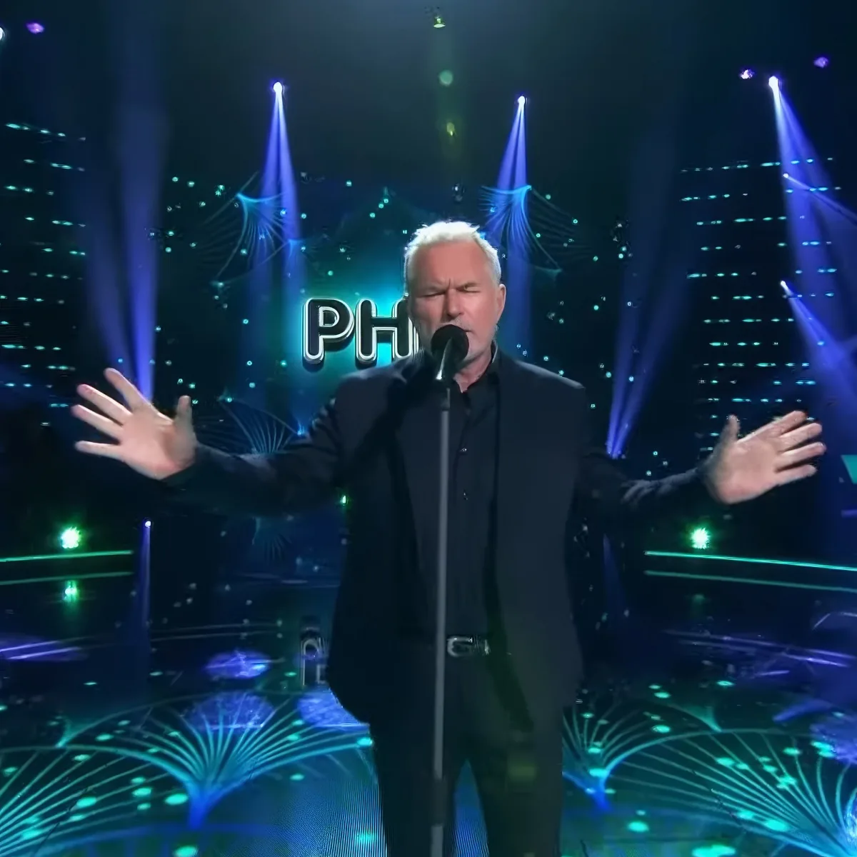 A man in black attire stands on stage with arms outstretched, microphone in front, under vibrant blue and green stage lights.