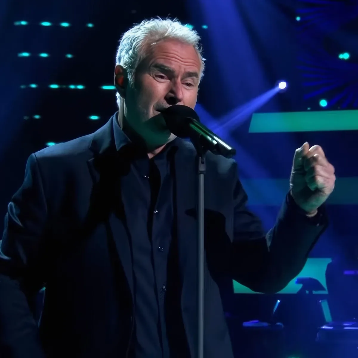 A man in a dark suit sings into a microphone on stage with blue lighting.