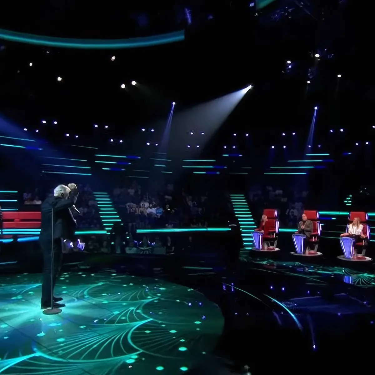 A performer in a dark suit stands on a stage with glowing green patterns, illuminated by spotlights.