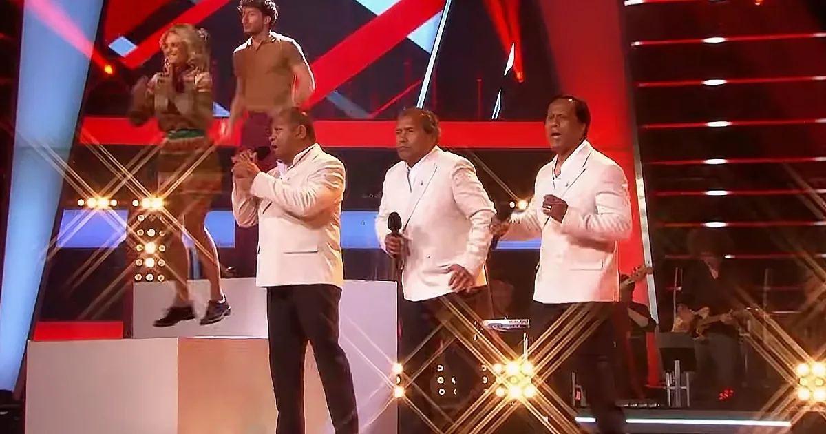 Three men in white suits sing on stage with a woman and man in the background.