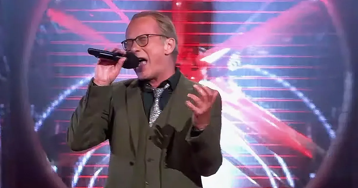 A man in a green suit sings into a microphone on a stage with colorful lights.