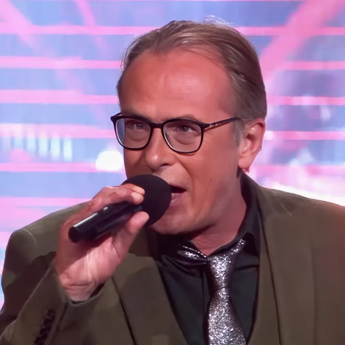 A man in a suit and bow tie speaks into a microphone against a colorful background.