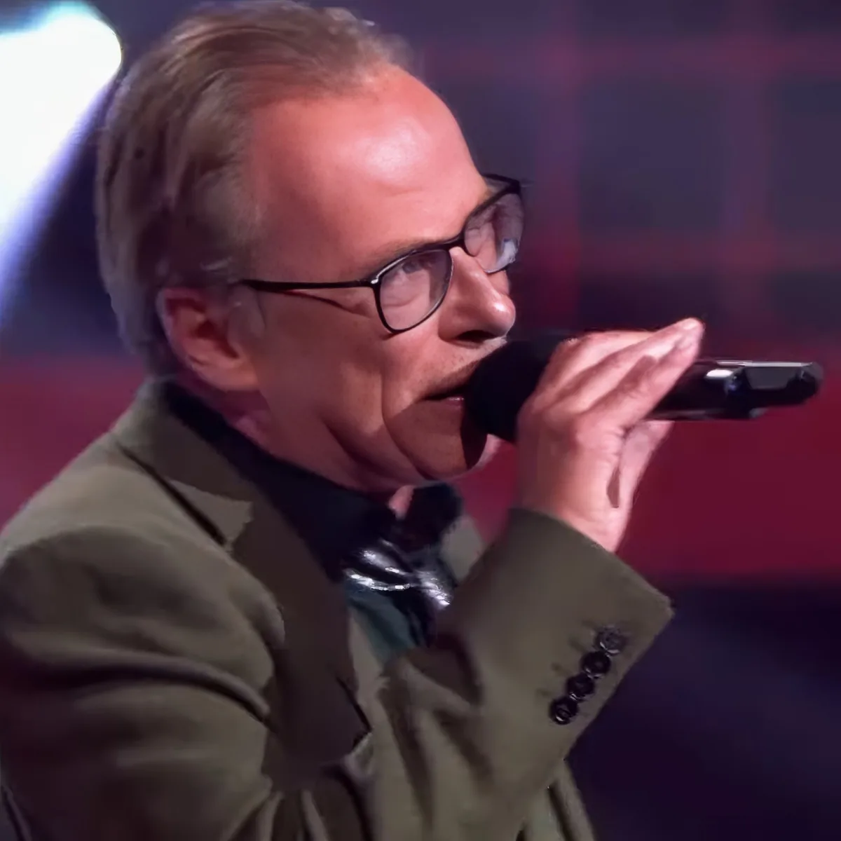 A man in a suit and glasses sings into a microphone on stage.