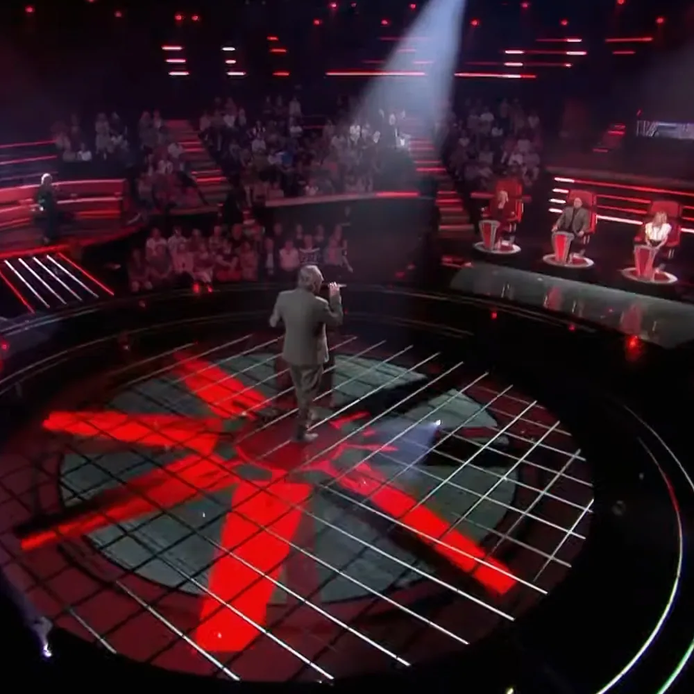 A man in a suit stands on a stage with a red and green grid floor, facing judges' chairs.