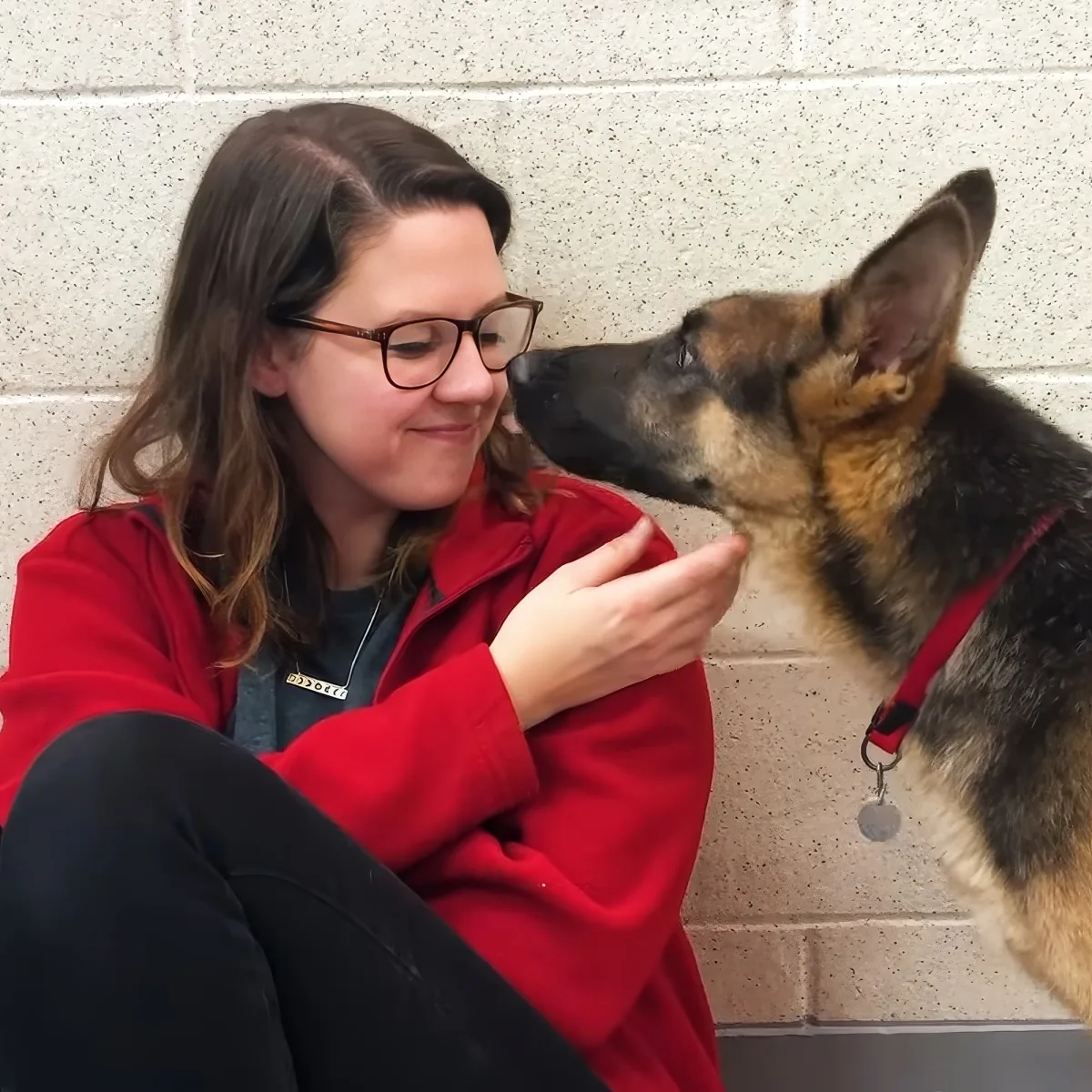 A woman in a red jacket pets a German Shepherd dog against a concrete wall.