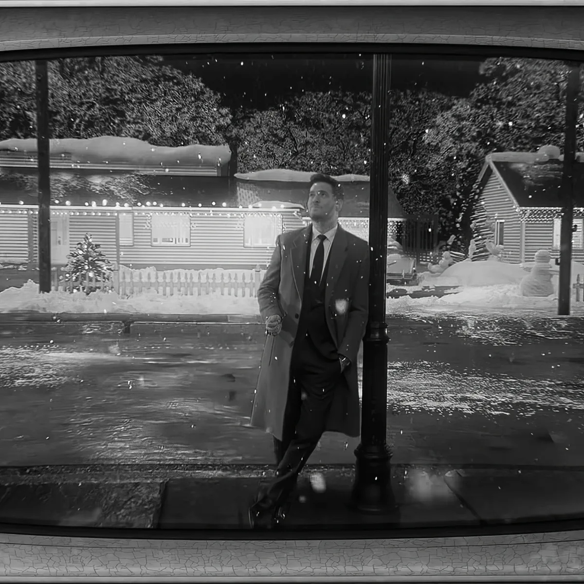 A man in a suit leans against a lamppost in a snowy, festive suburban scene.