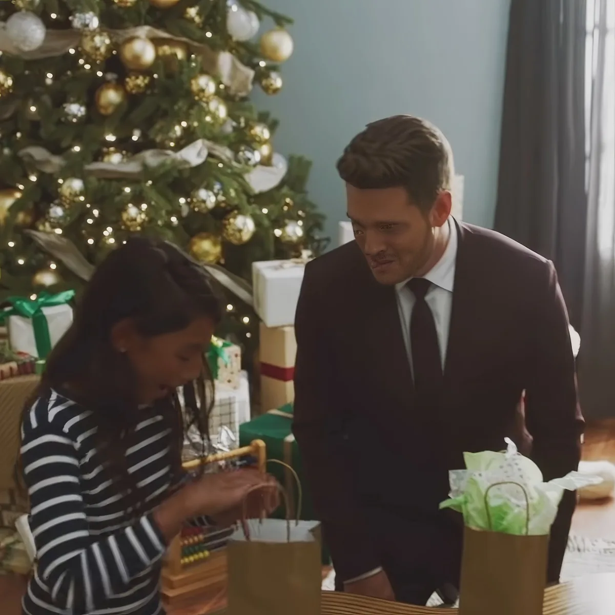 A man in a suit and a young girl in a striped dress are near a Christmas tree, surrounded by gifts.