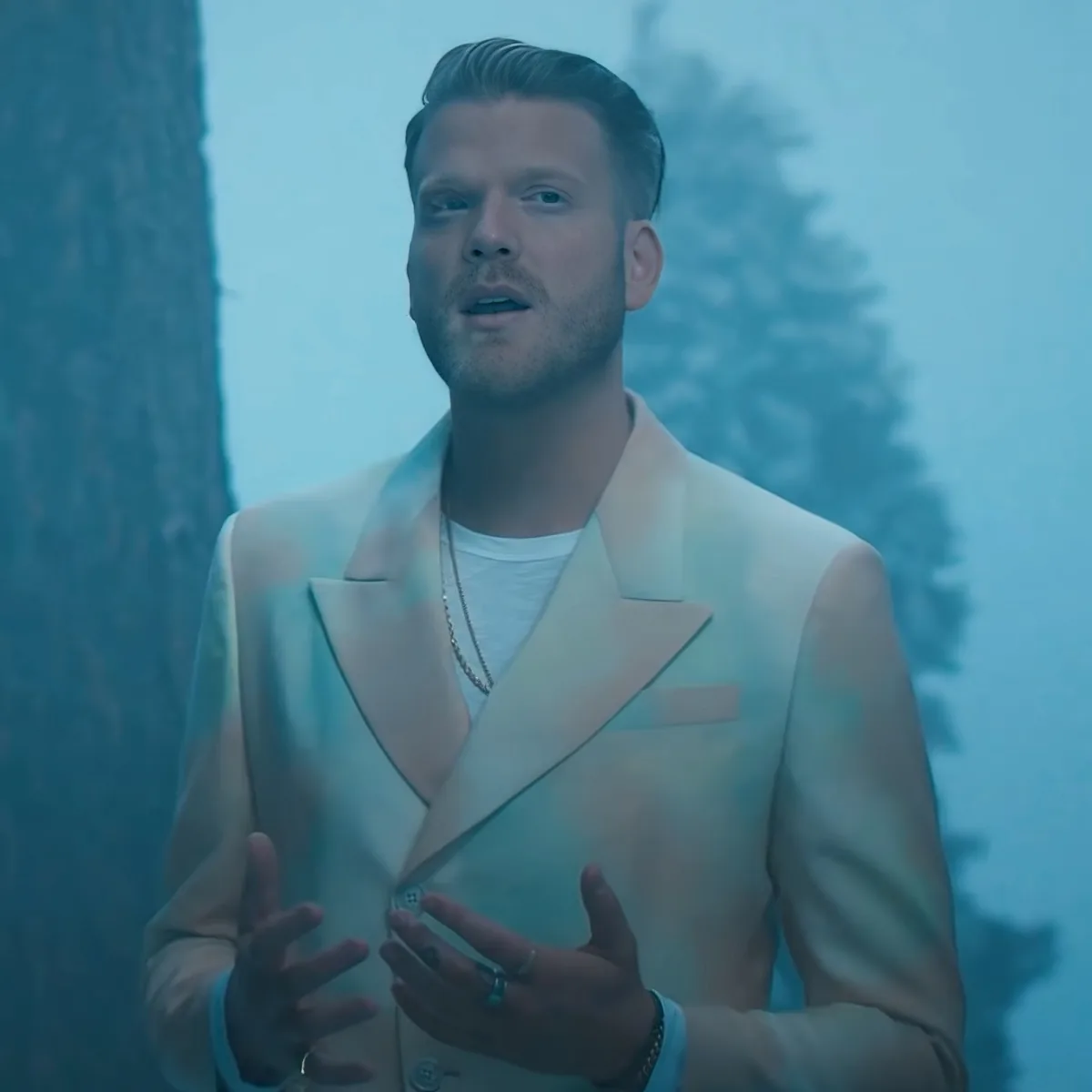 A man in a light-colored suit jacket and white shirt stands outdoors, gesturing with his hands.