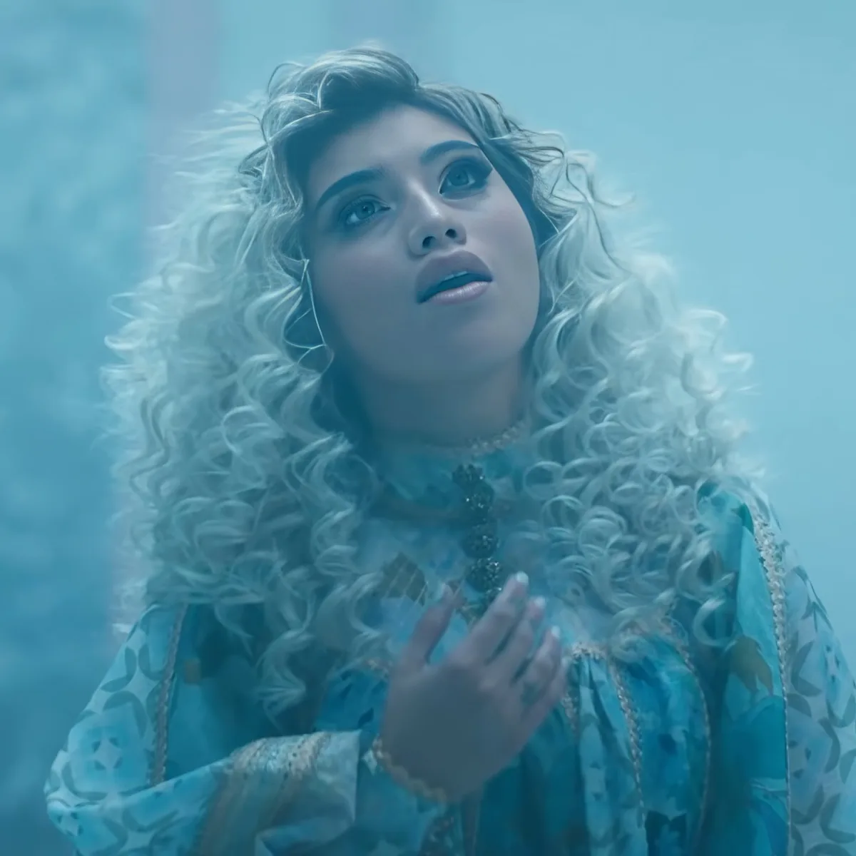 A person with long, curly white hair and a blue patterned outfit is holding their hands together.