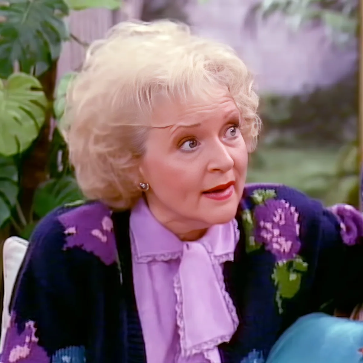An elderly woman with blonde hair, wearing a purple floral sweater and a pink blouse, sits in a garden setting.