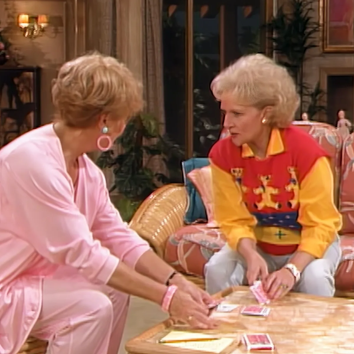 Two women in a cozy room, one in pink, the other in colorful attire, are engaged in a card game at a wooden table.