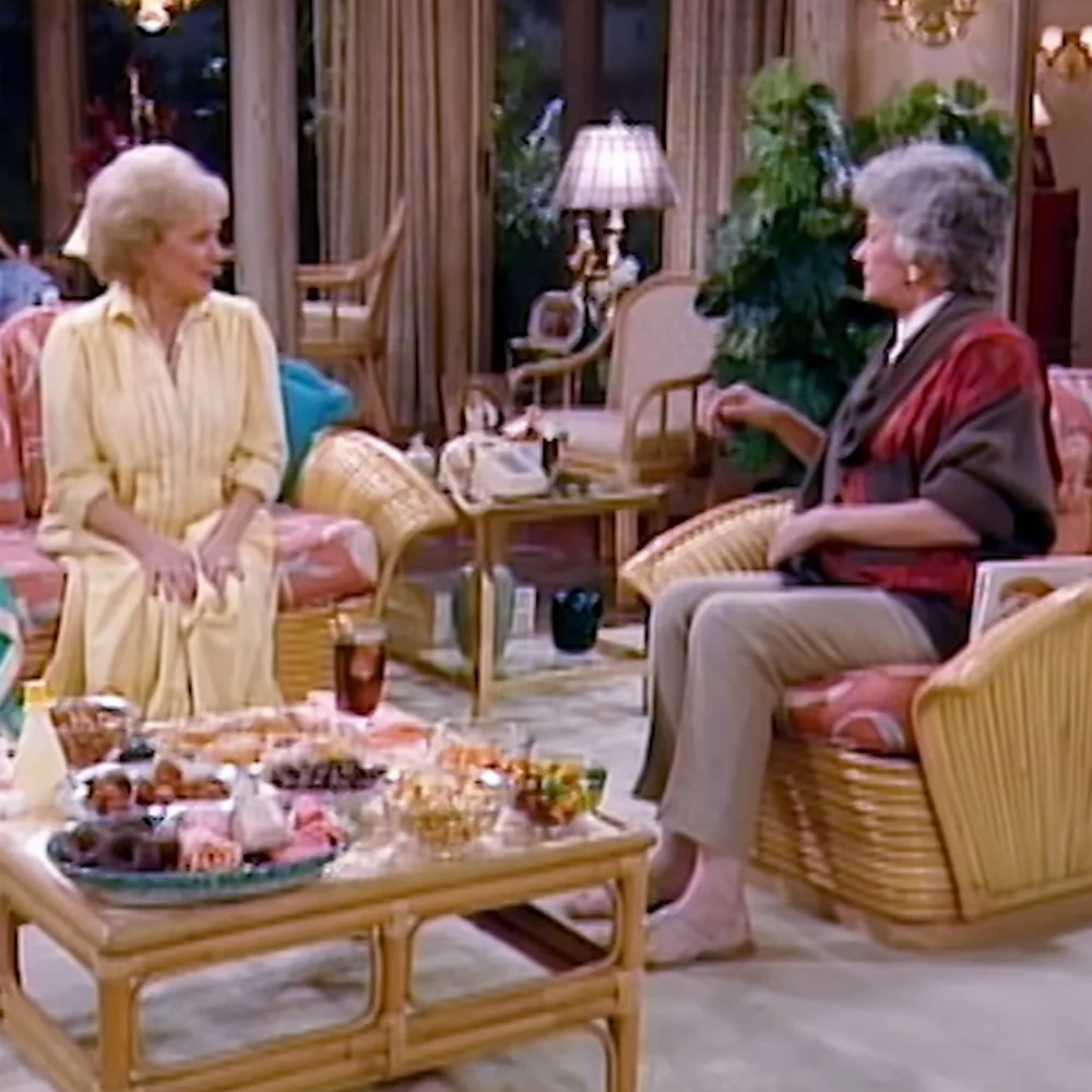 Two women are seated in a cozy living room, engaged in conversation, surrounded by wicker furniture and a table with...