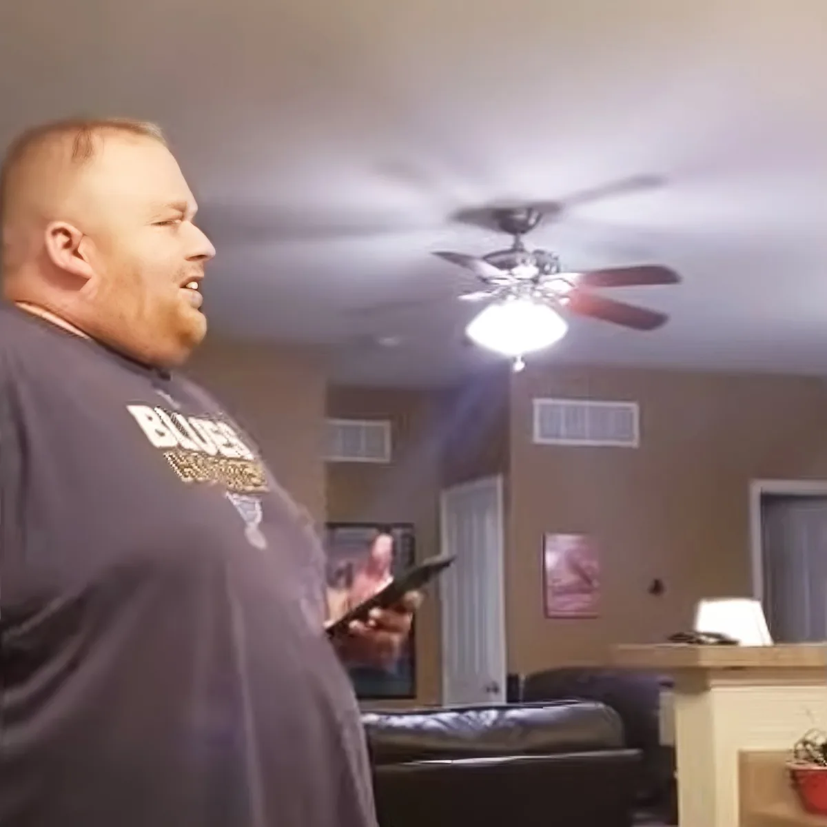 A man in a "Blues" shirt holds a phone, looking up at a ceiling fan in a room with a couch and lamp.