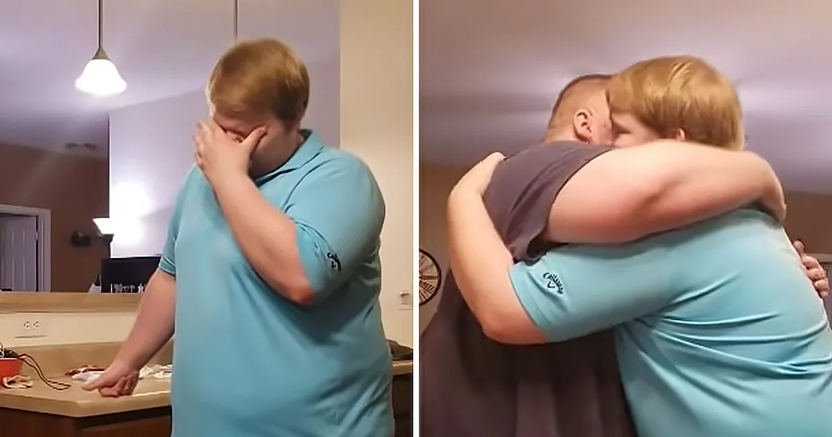 A person in a blue shirt is embracing another person in a dark shirt in a kitchen setting.