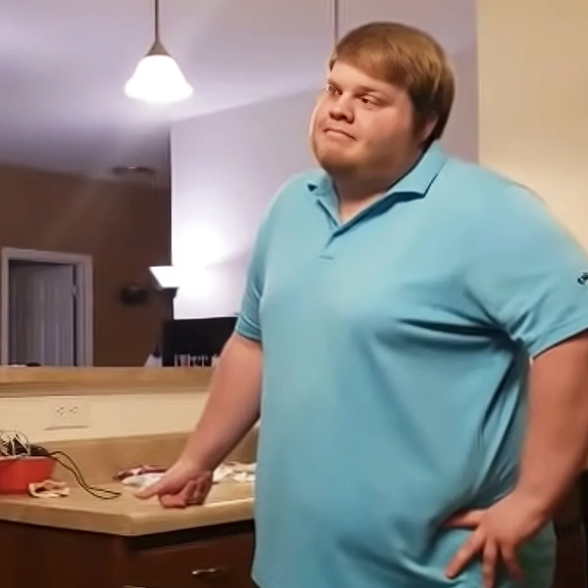 A person in a light blue polo shirt stands in a kitchen, hands on hips, looking to the side.