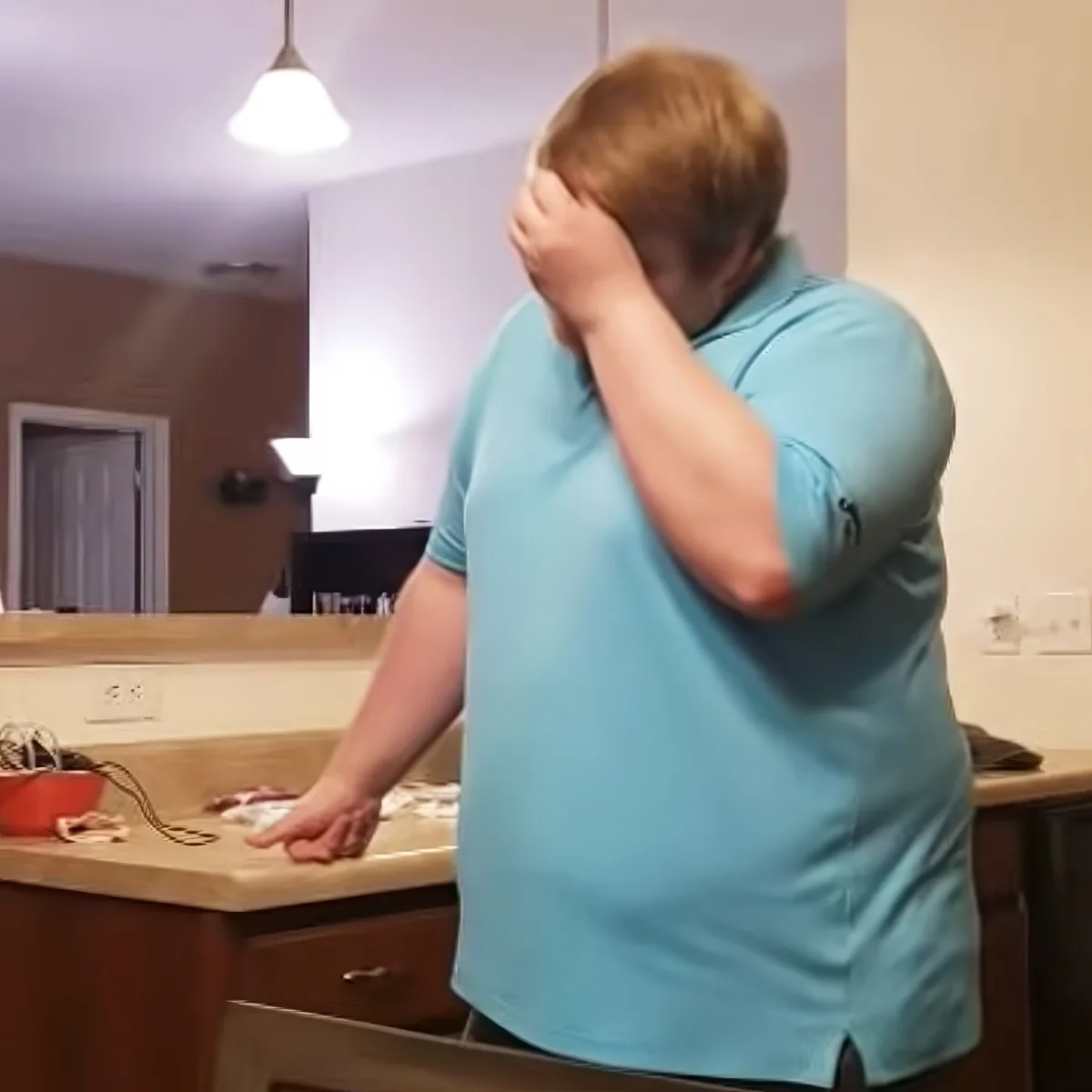 A person in a light blue shirt stands in a kitchen, covering their face with one hand and reaching out with the other.