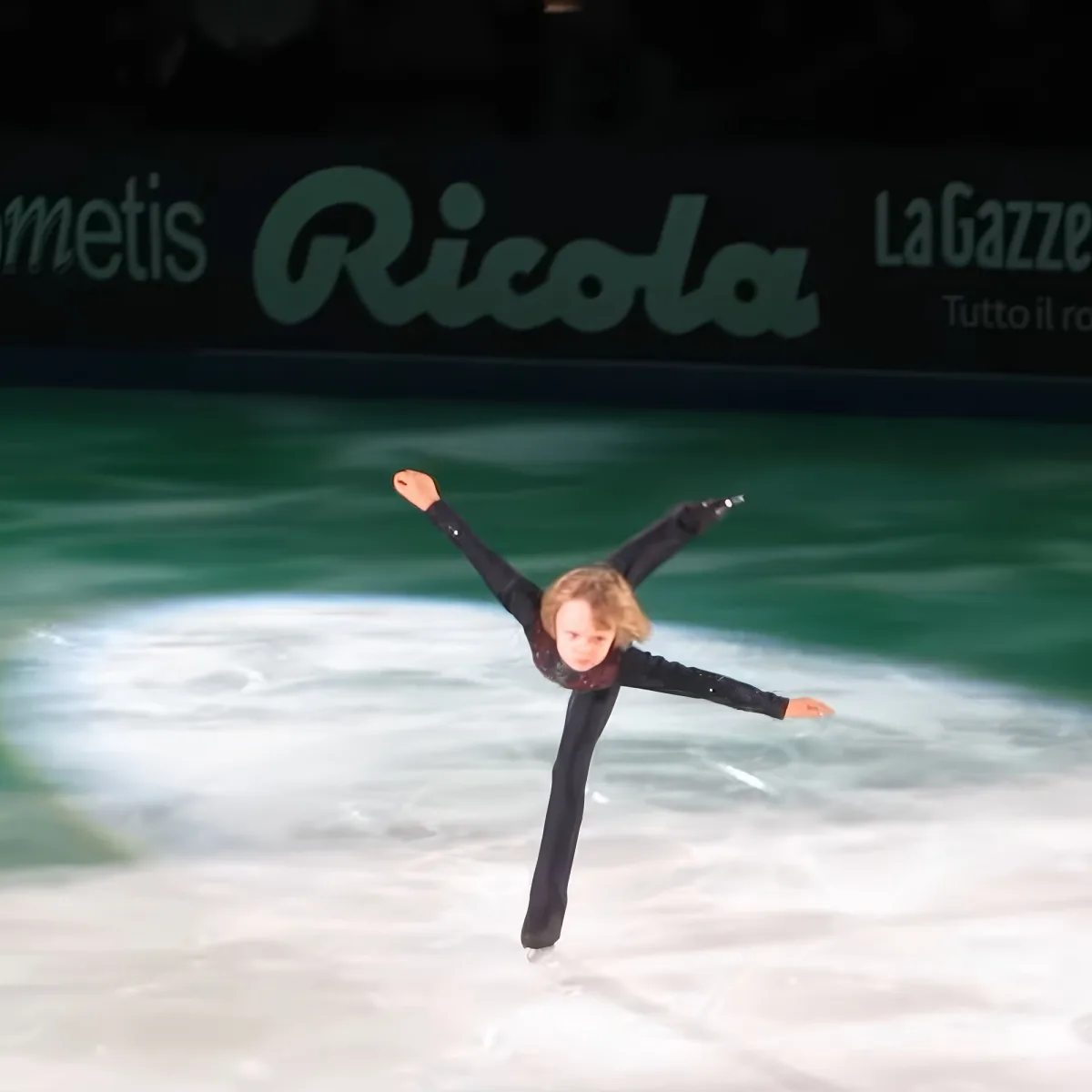 A young figure skater in a black outfit performs a split jump on ice.