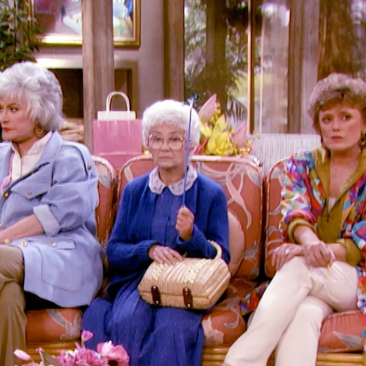 An elderly woman in a blue sweater sits on a couch holding an umbrella and a handbag.