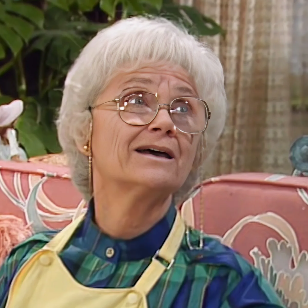An elderly woman with white hair and glasses, wearing a green and blue striped shirt and a yellow apron, sits on a...