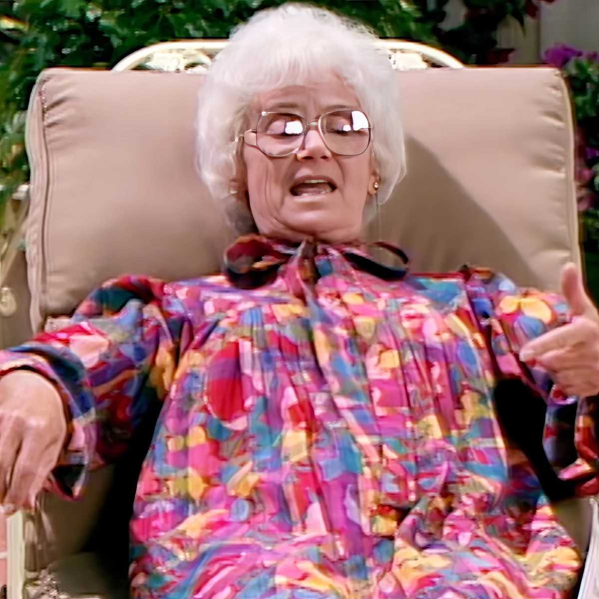 An elderly woman with white hair, wearing glasses and a colorful dress, sits in a chair with her arms outstretched.
