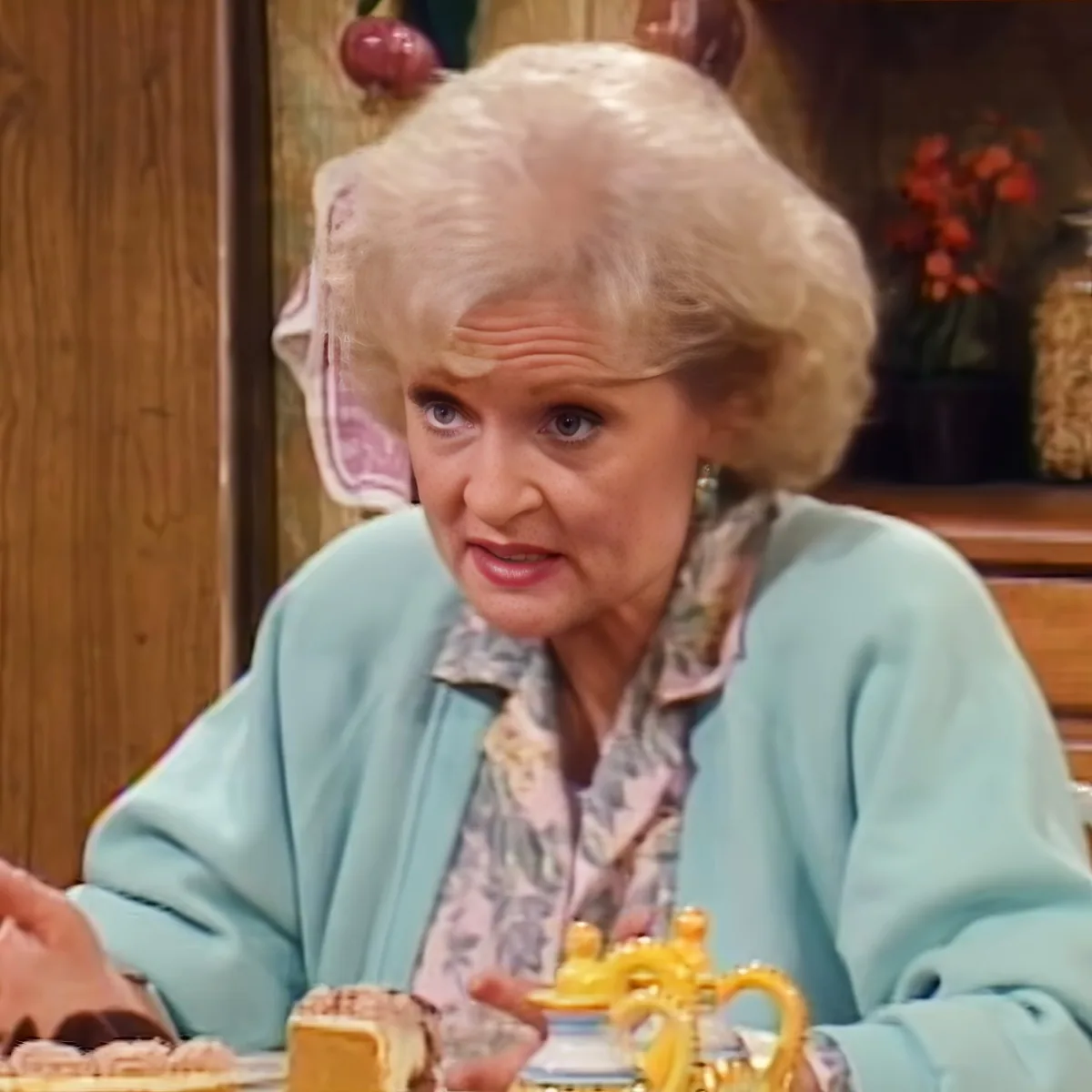 An older woman with blonde hair, wearing a light blue sweater and floral shirt, sits at a table with a teapot and cake in...
