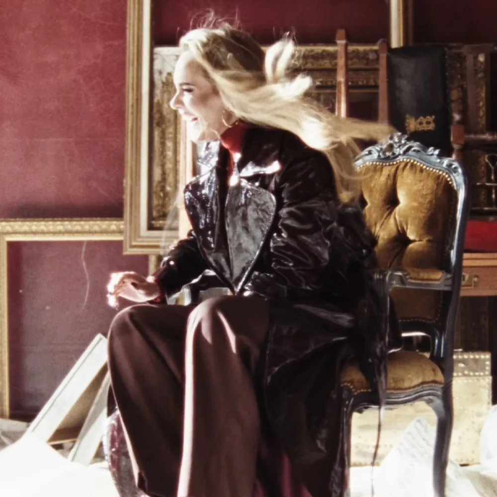 A woman in a black leather jacket sits on an ornate chair, her hair blowing in the wind.
