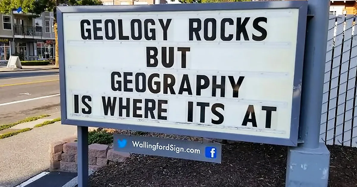 A sign humorously states "Geology rocks but Geography is where its at" in a suburban setting.