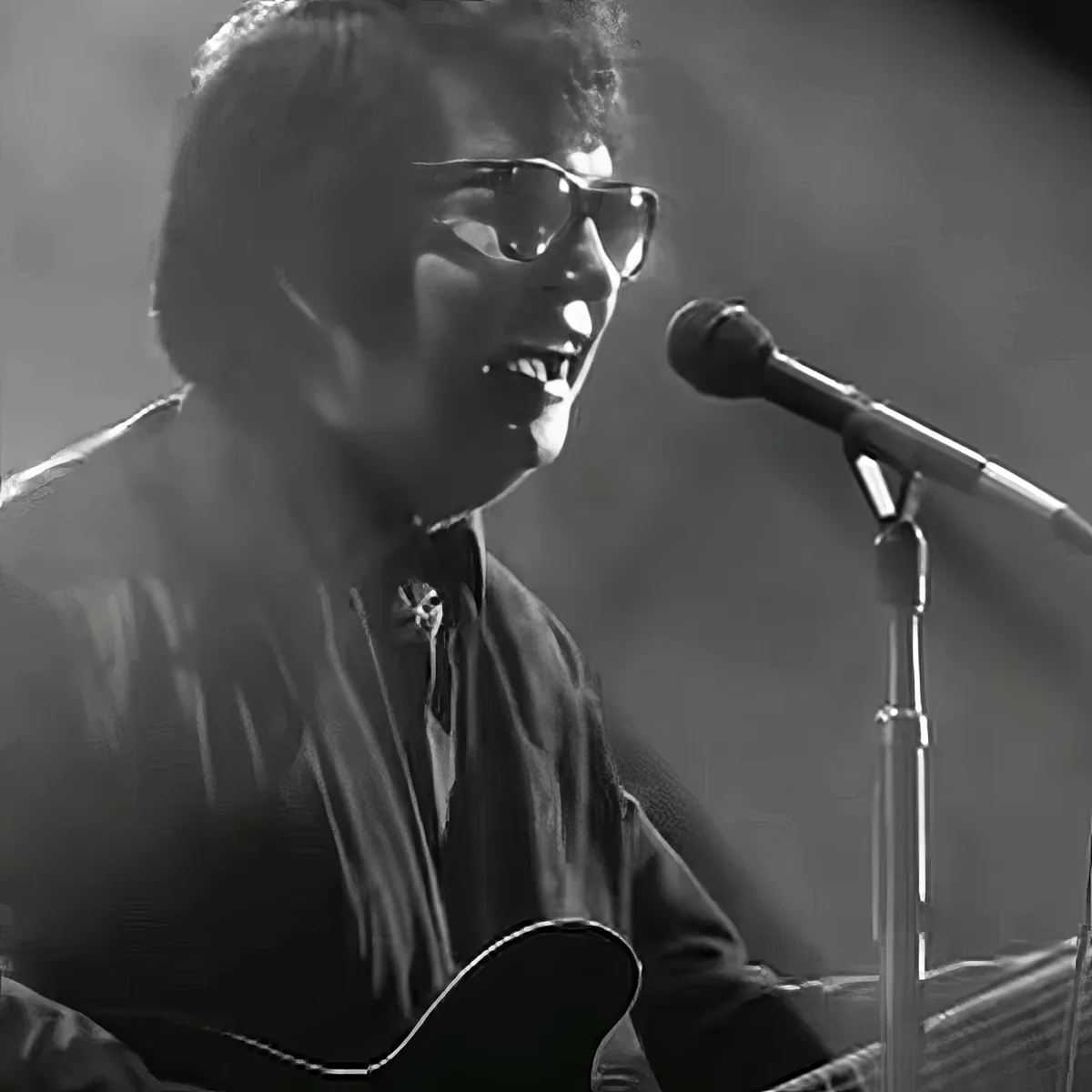 A person wearing glasses and a striped shirt sings into a microphone while holding a guitar.