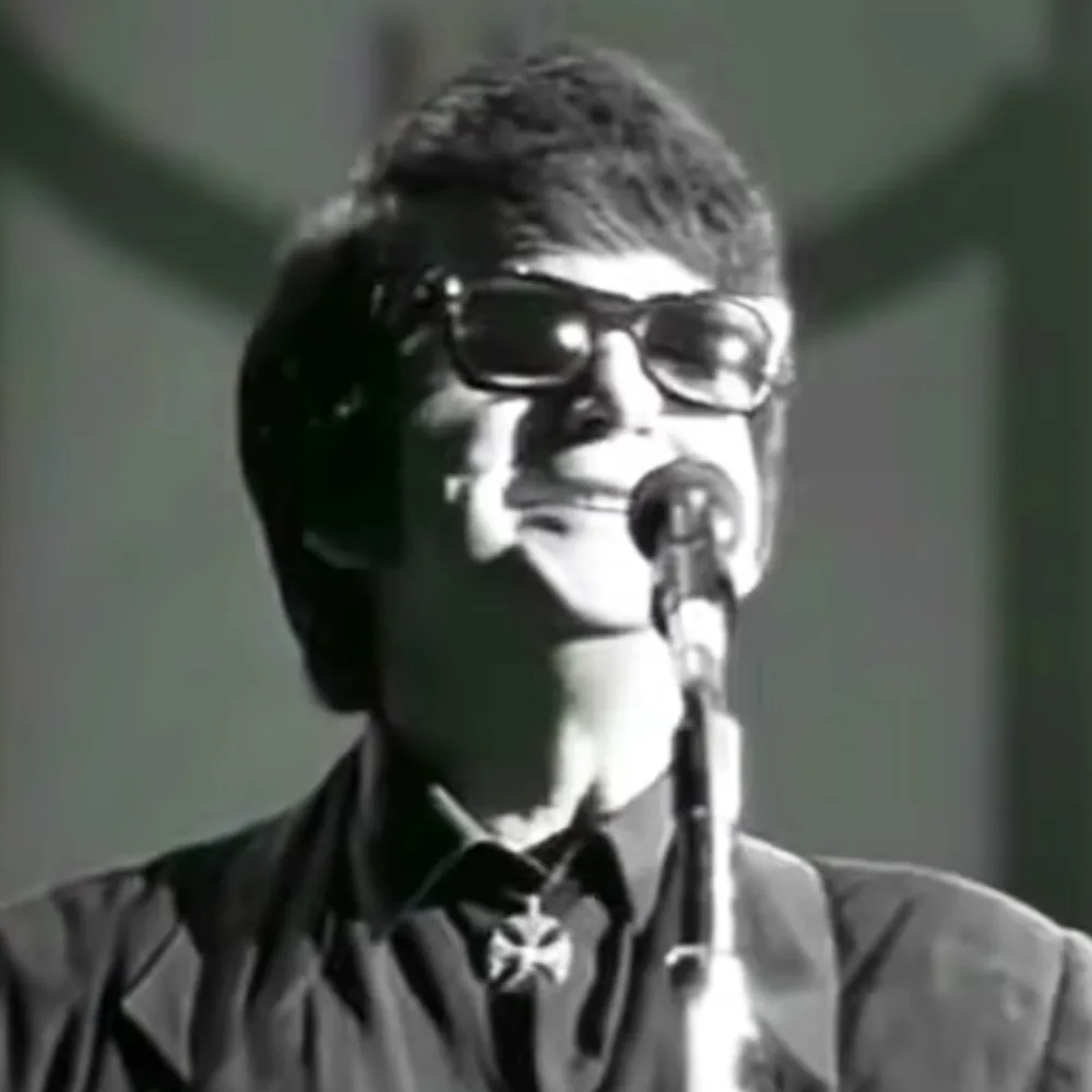A person wearing glasses and a tie stands in front of a microphone, possibly singing or speaking.