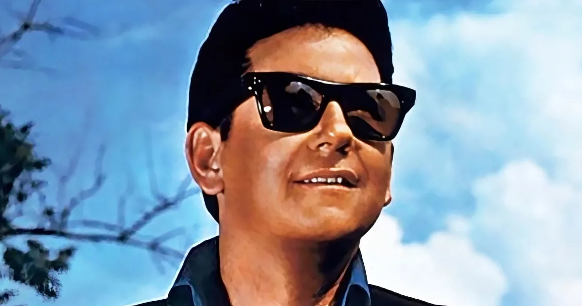 A person wearing sunglasses and a dark cap smiles against a blue sky with clouds and tree branches.