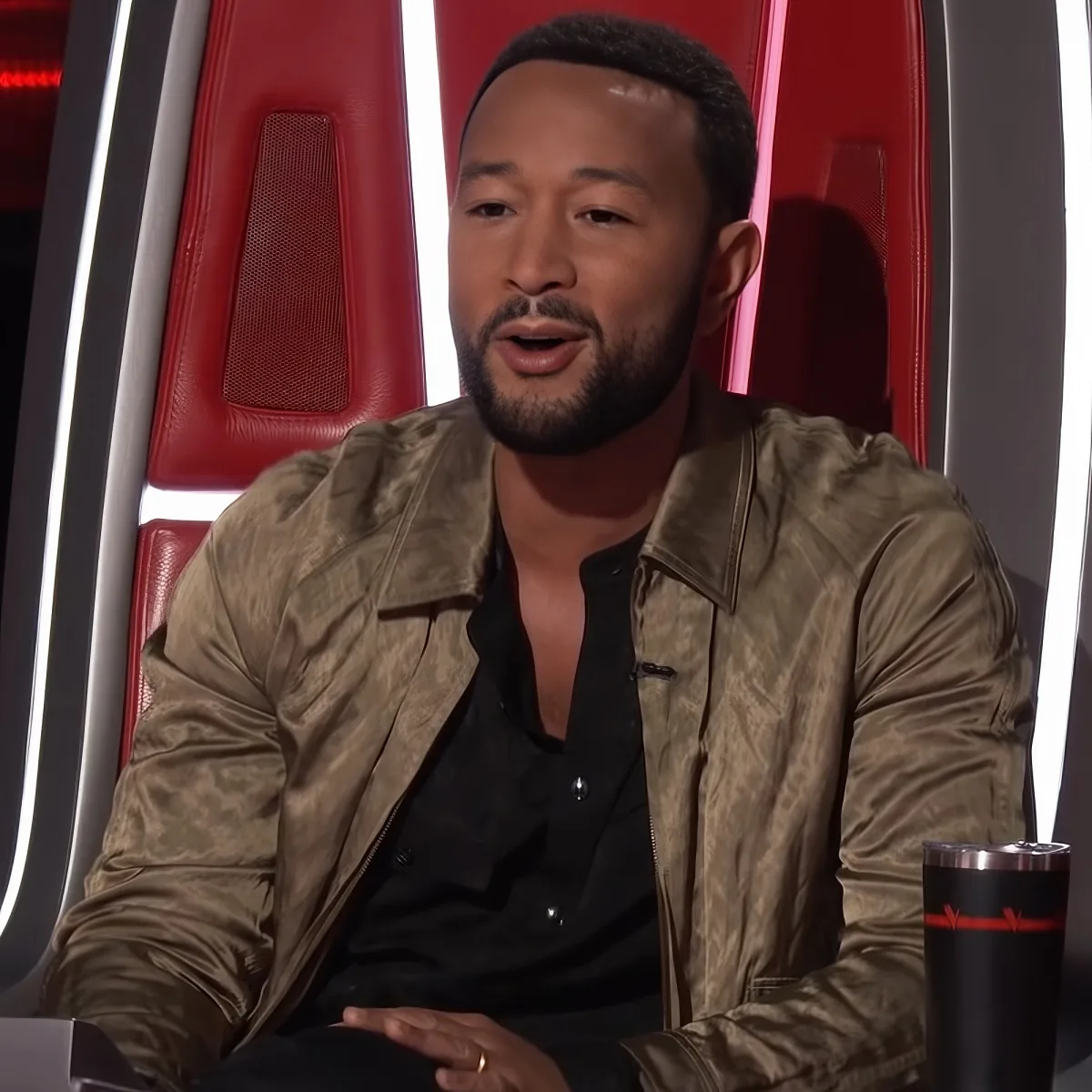 A man with a beard, wearing a gold jacket and black shirt, sits in a red chair, appearing to be on a talk show or interview.