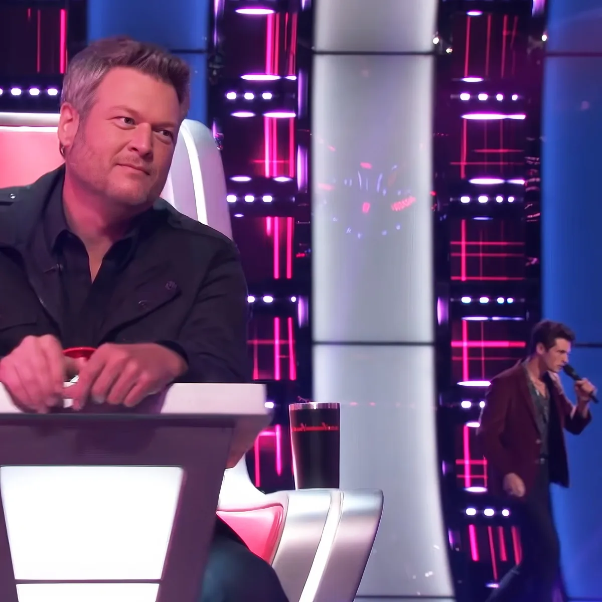 A man in a black suit sits in a chair, looking to the side, with another man singing on stage in the background.
