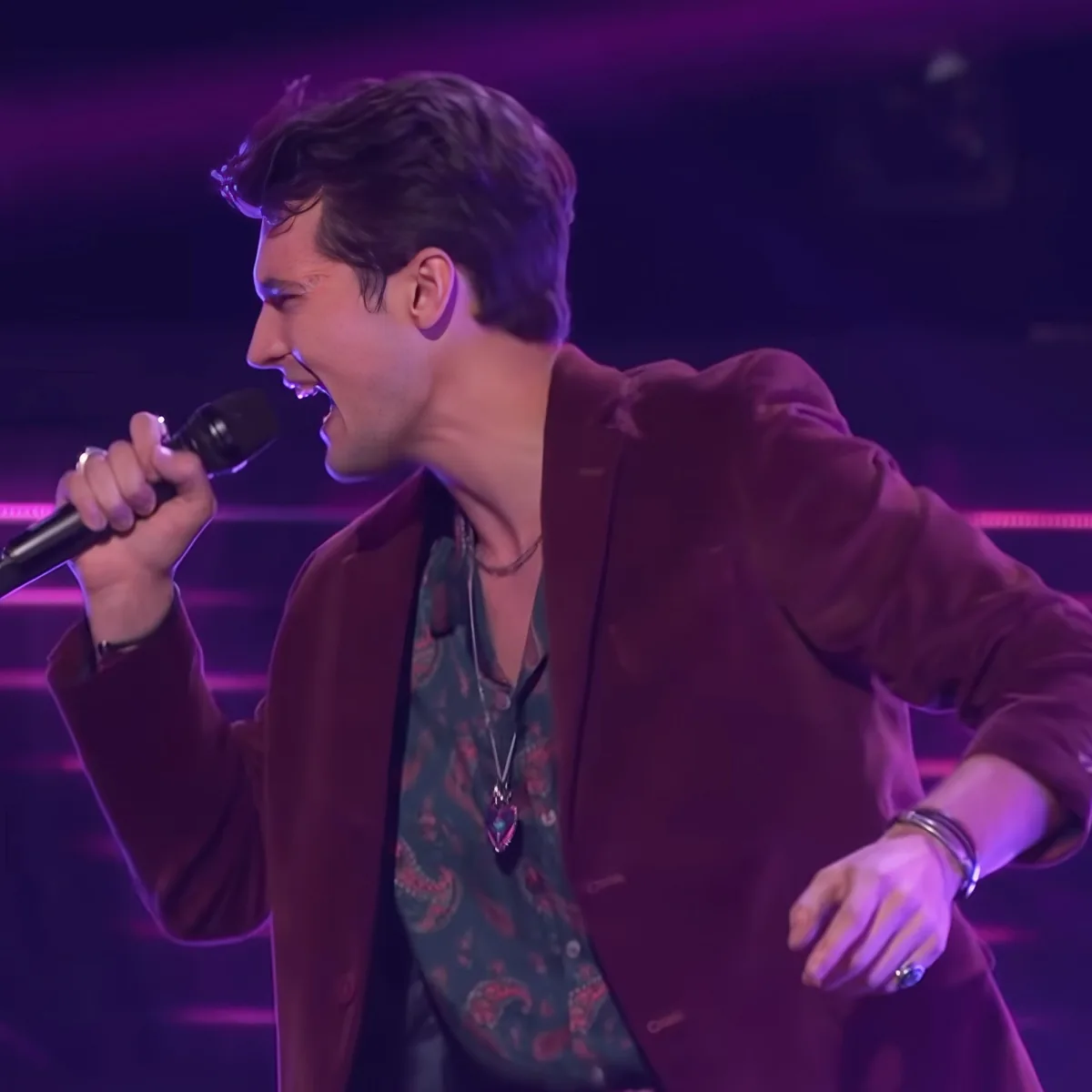 A man in a maroon blazer sings passionately into a microphone on stage.