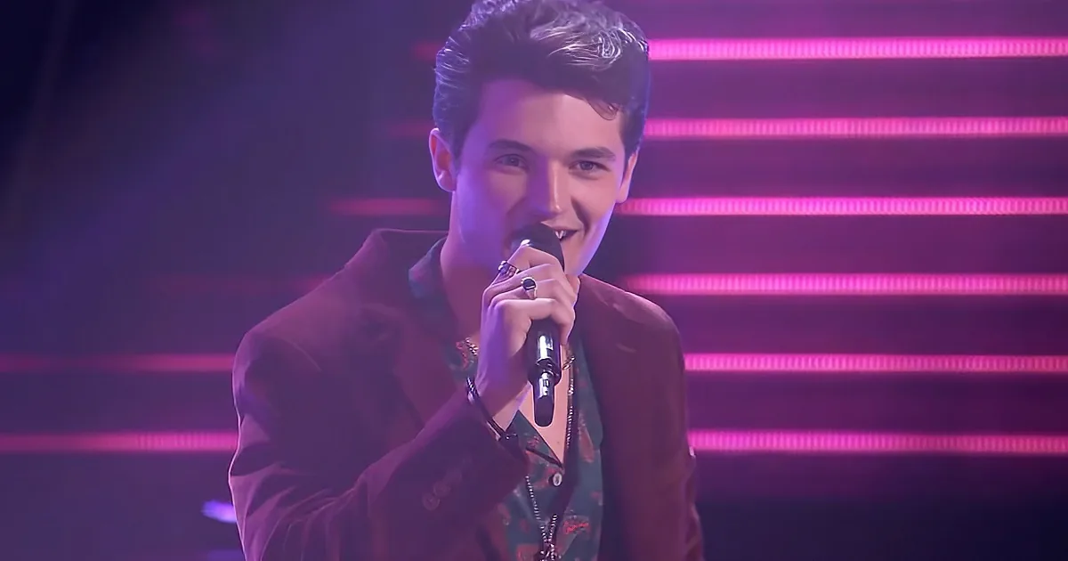 A person in a dark suit holds a microphone, wearing rings, against a backdrop of pink and purple lights.