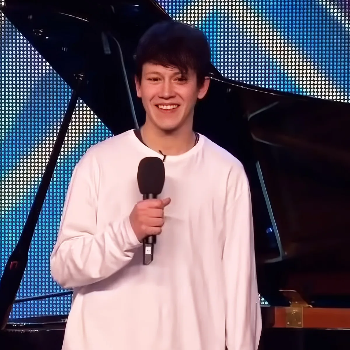 A person in a white shirt holds a microphone, smiling, with a piano and blue LED lights in the background.
