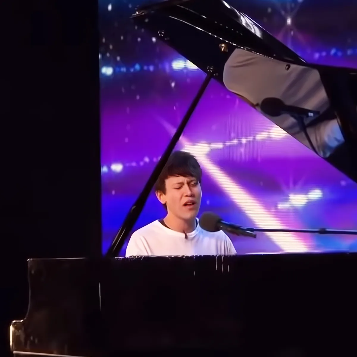 A person in a white shirt sings into a microphone while playing a grand piano on a stage with purple lighting.