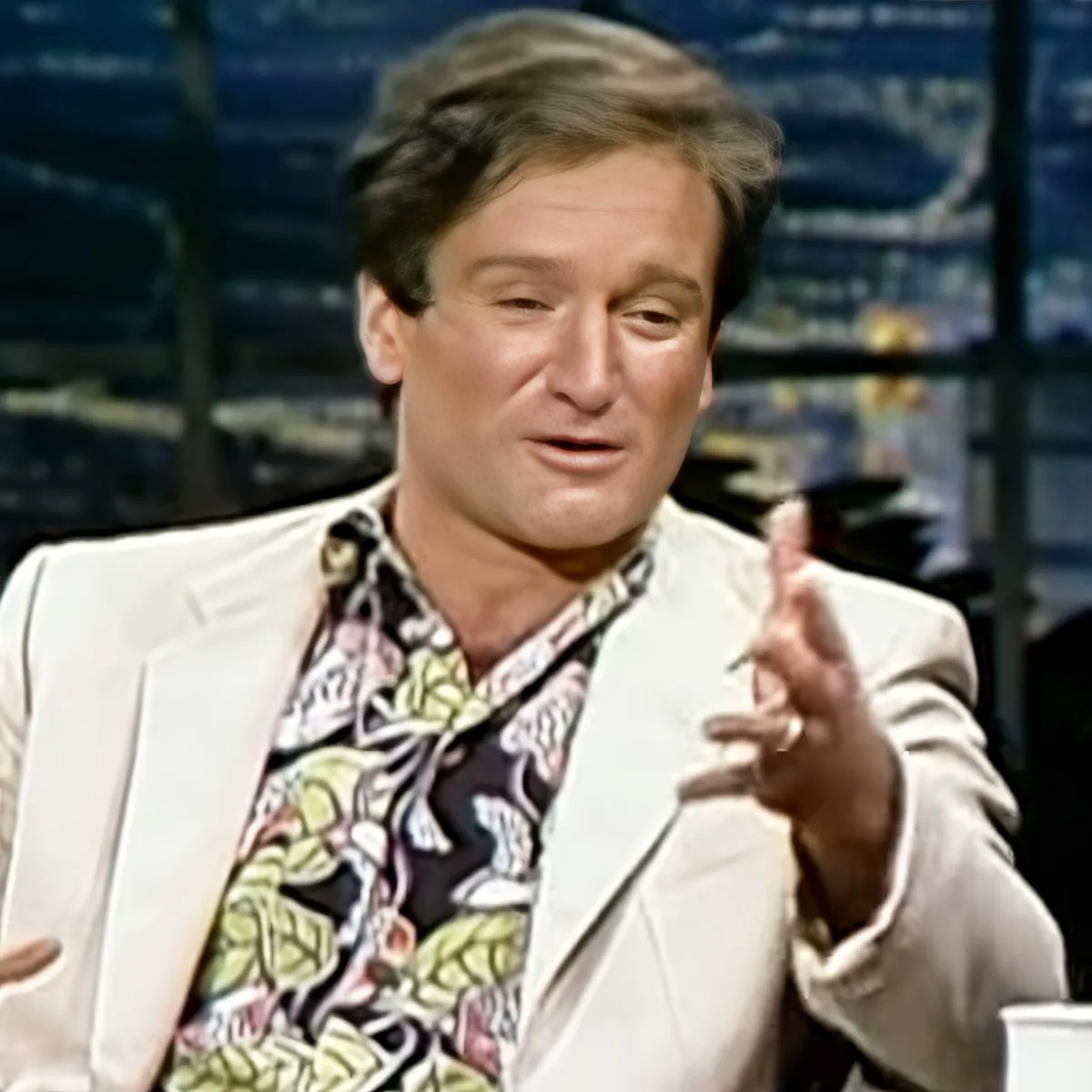 A man in a white blazer and colorful shirt gestures while speaking, possibly on a talk show set.