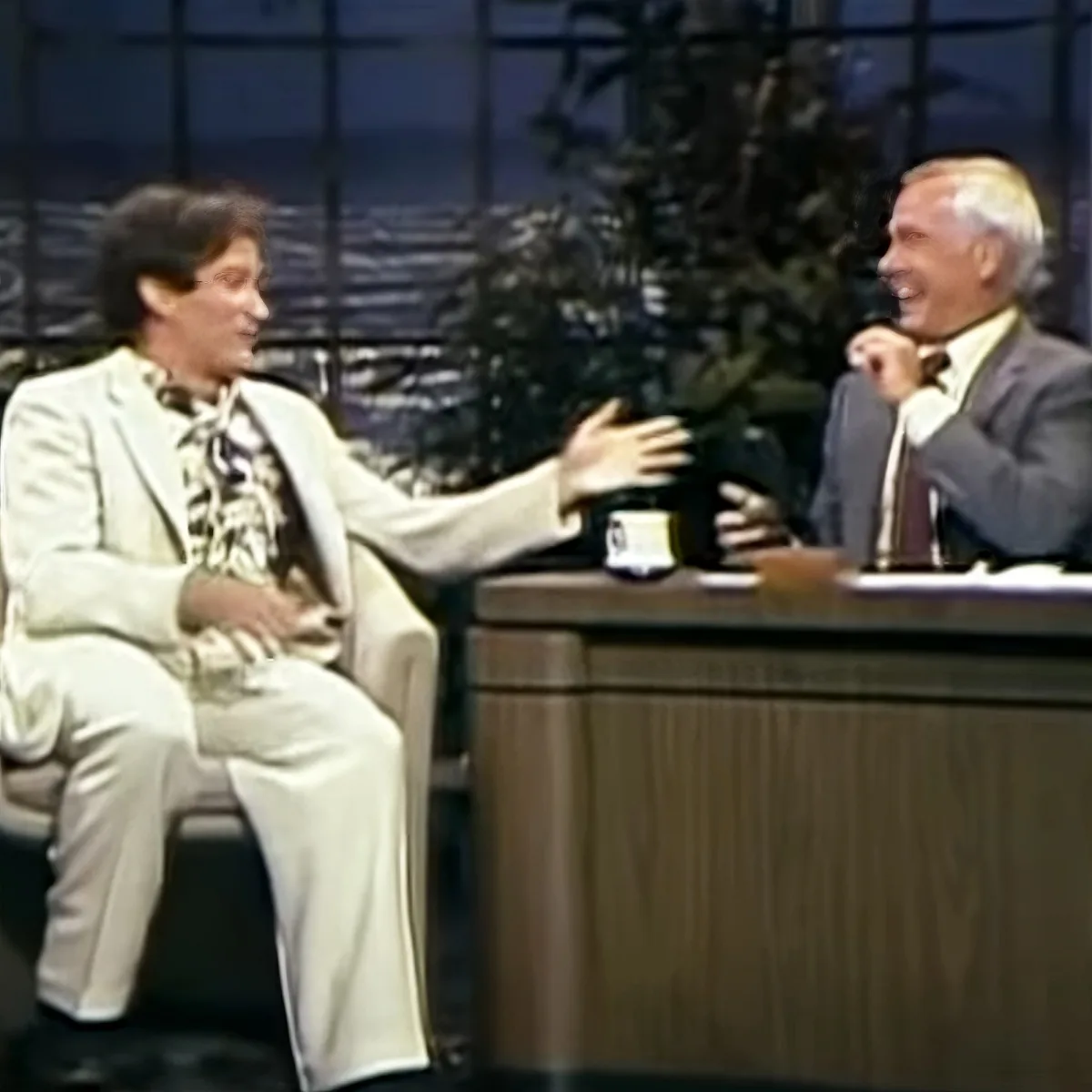 A man in a white suit extends his hand towards another man in a suit, both seated, in a studio setting.