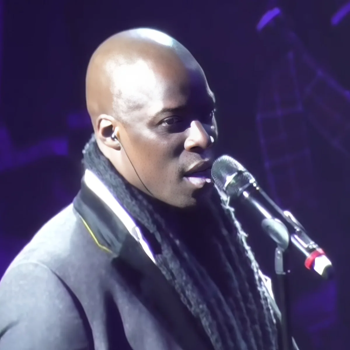 A man with a bald head and earphones sings into a microphone on stage.