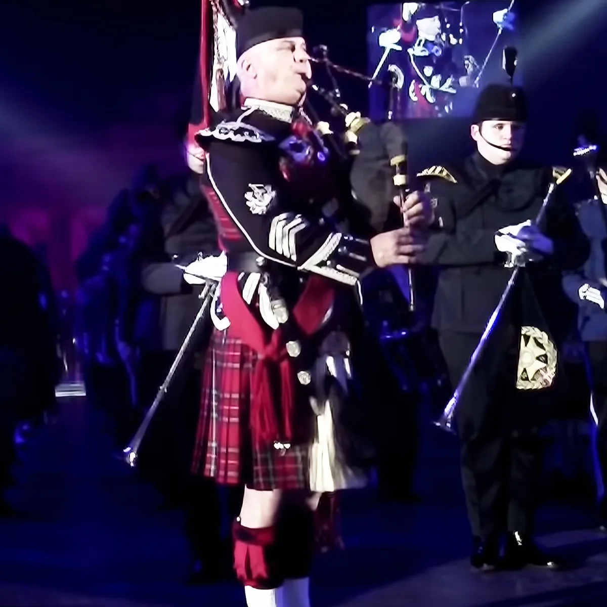 A man in a kilt and military uniform plays bagpipes on stage, surrounded by other uniformed individuals.
