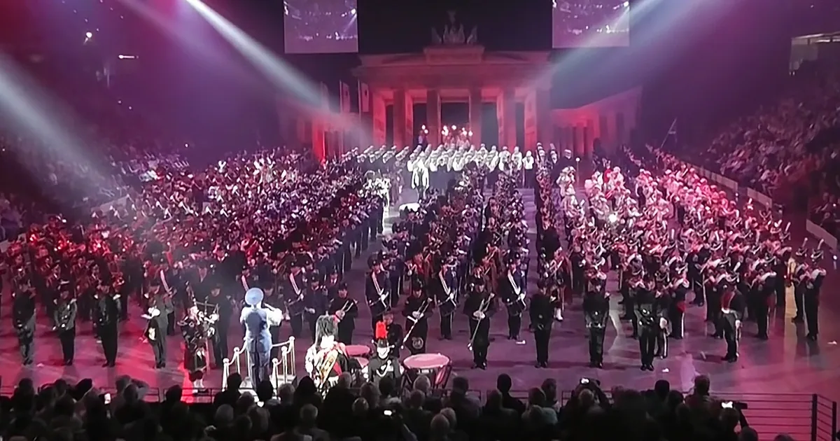 A marching band in formal attire performs on stage with a large crowd and illuminated backdrop.