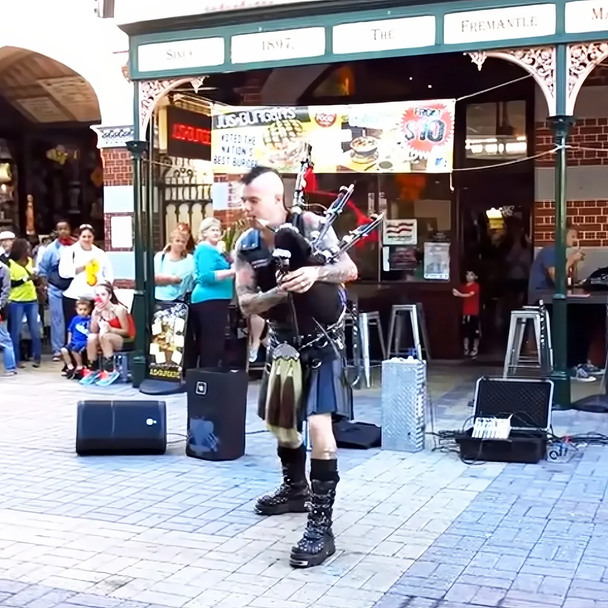 A man in a kilt plays bagpipes on a street, surrounded by people and speakers.