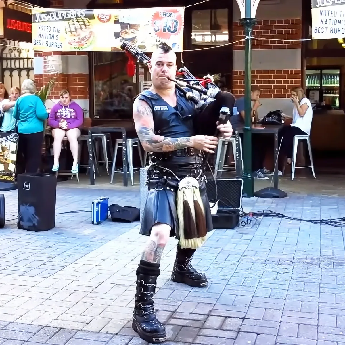 A man in a kilt plays bagpipes on a brick street, surrounded by people and a sign for "Jus Burgers."