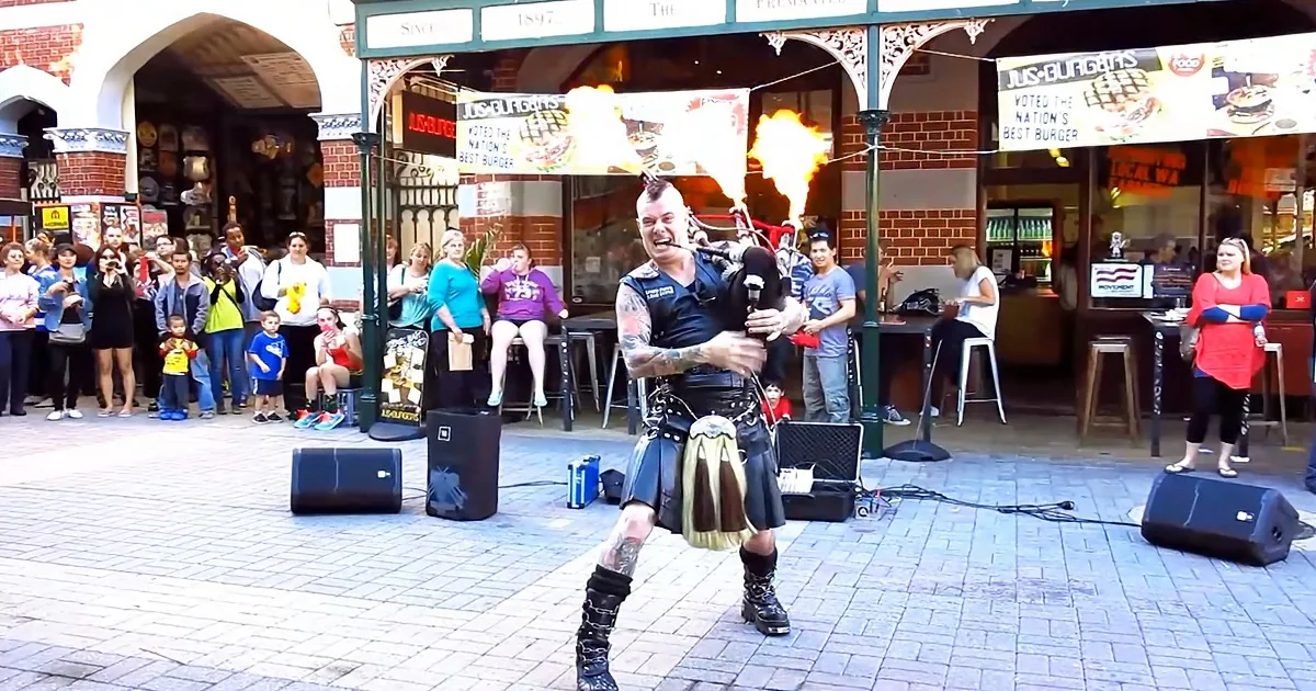 A man in a kilt plays bagpipes while flames shoot from his mouth, captivating a crowd in a bustling outdoor market.